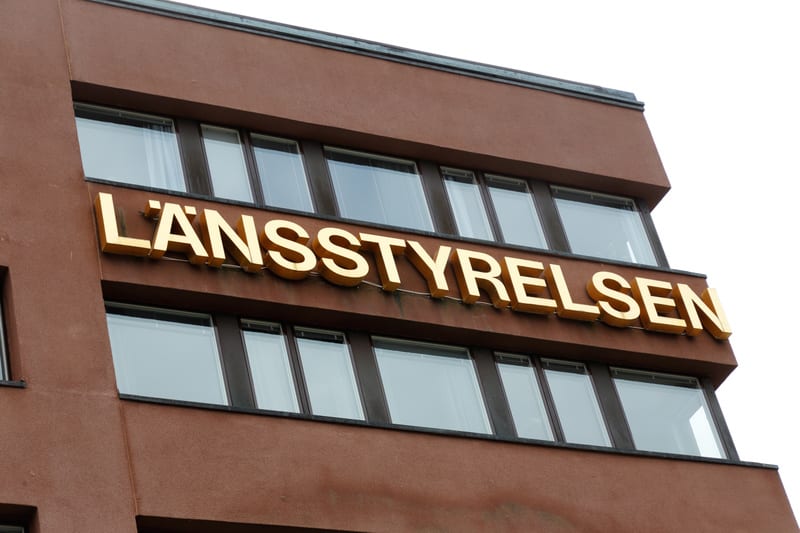 Bilden visar fasaden på en brun byggnad under en klar, vit himmel. Ett stort guldgult skylt med texten "LÄNSSTYRELSEN" är monterat på fasaden, mellan två rader av fönster. Fönstren är rektangulära med mörka ramar och reflekterar ljuset. Byggnadens hörn, där skärmen sitter, är vinklat, vilket ger byggnaden en modern arkitektur. Fasadytan verkar vara i sten eller putsade tegel.