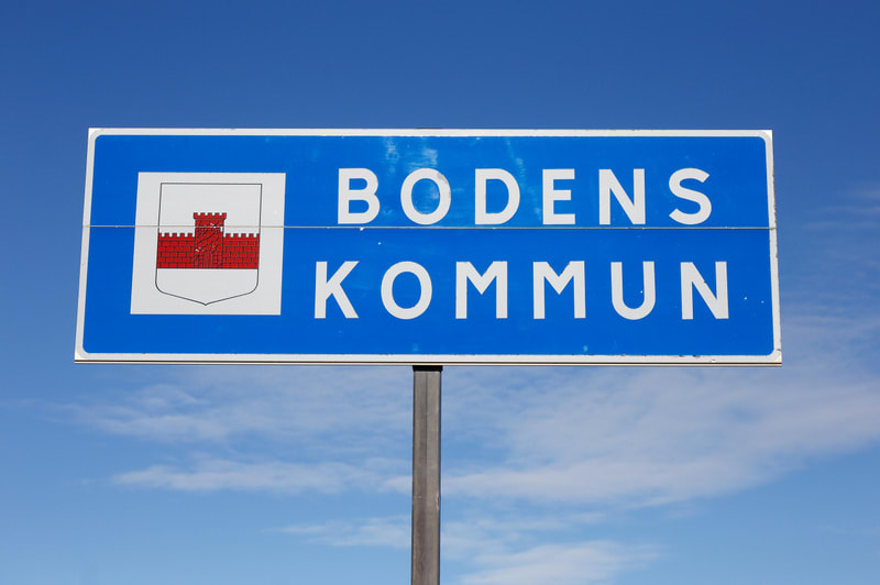 En blå skylt med vit text som säger "BODENS KOMMUN" och en vapensköld syns framför en klarblå himmel med några vita moln. Skylten är rektangulär och monterad på en grå stolpe som sticker upp nedanför skylten. Vapenskölden på skyltens vänstra sida är