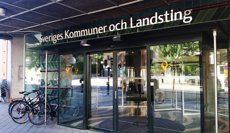 Bilden visar entrén till en byggnad med texten "Sveriges Kommuner och Landsting" ovanför de automatiska glasdörrarna. Till vänster om entrén står två cyklar parkerade. Entrédörrarna reflekterar omgivningen, inklusive träd, en gata med trafikljus och vad som ser ut att vara en annan byggnad. En skylt med en gul och röd logotyp syns reflekterad i glasdörrarna. Utanför dörrarna, på marken, finns en mörkare matta/golvdekoration med ett vågmönster. Taket över entrén är mörkt med inbyggd belysning.