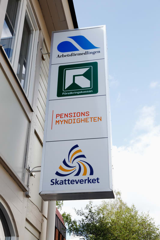 Bilden visar en vertikal skylt fäst på en byggnadsfasad. Skylten innehåller logotyper och namn för fyra svenska myndigheter: Arbetsförmedlingen, Försäkringskassan, Pensionsmyndigheten och Skatteverket. Ovanför skylten syns en del av en vit träfasad med ett fönster. Till höger om skylten syns en ljusblå himmel med några vita moln, samt toppen av ett träd med gröna löv i nedre höger hörn.