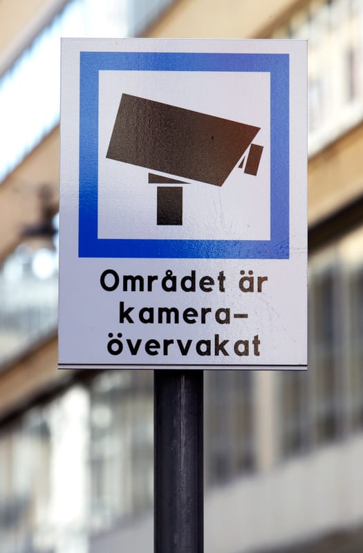 En vertikal bild visar en skylt med texten "Området är kameraövervakat" på svenska. Skylten är vit med en blå ram runt en svart kamerasymbol i överkanten. Skylten är monterad på en svart stolpe. I bakgrunden syns suddiga byggnader i ljusa färger, vilket indikerar att bilden är tagen utomhus under dagtid. Färgerna på byggnaderna i bakgrunden är främst beige och gråaktiga.