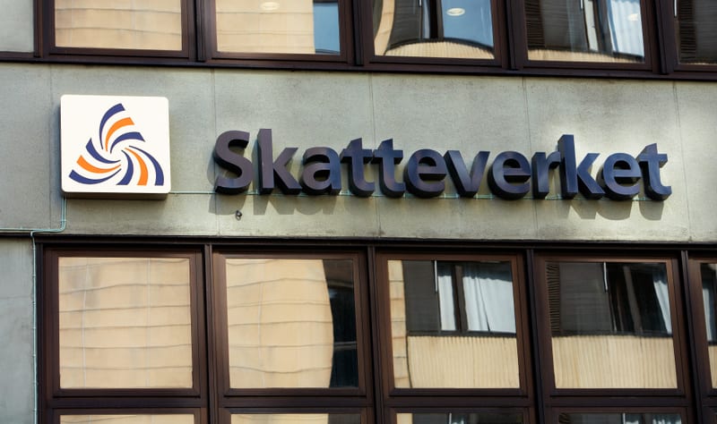 Bilden visar fasaden på en byggnad, troligtvis under dagtid. Centralt i bilden syns en skylt med texten "Skatteverket" i mörkare, upphöjda bokstäver. Till vänster om texten finns en fyrkantig logotyp med en stiliserad virvel i blått och orange. Fasaden verkar vara av en ljusare sten- eller betongtyp. Nedanför skylten och logotypen löper en rad med fönster med mörkare ramar. I fönsterglasen reflekteras omgivningen, inklusive delar av en annan byggnad och eventuellt gardiner eller persienner på insidan.