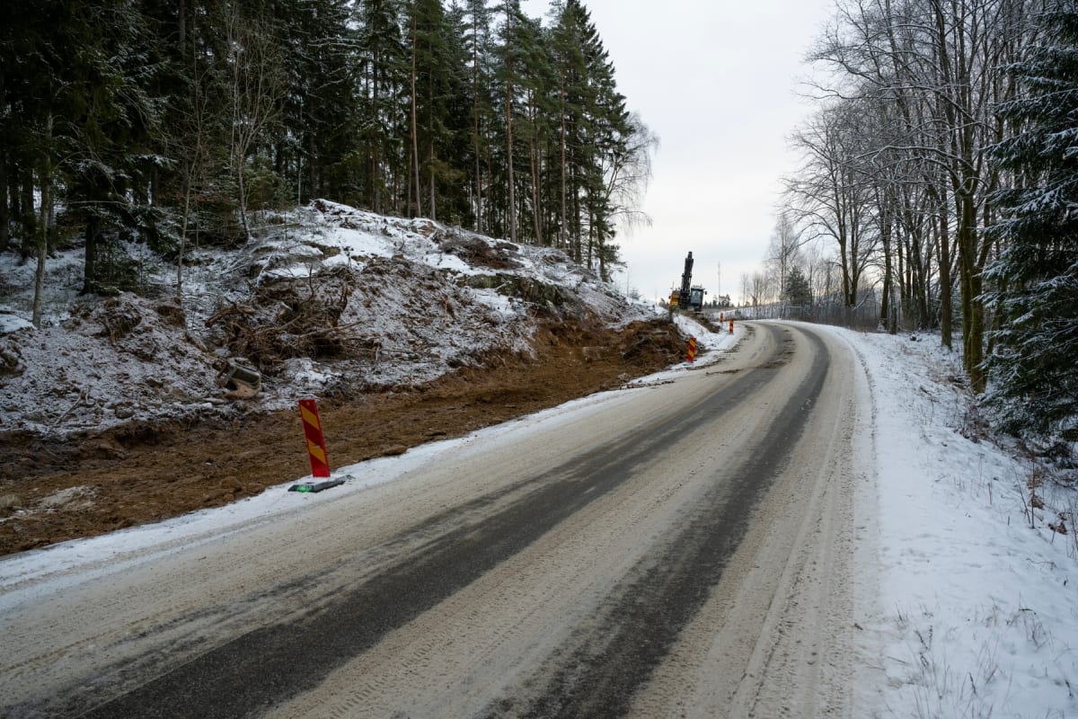 Bilden visar en snötäckt grusväg som svänger åt höger, omgiven av skog och snötäckt mark. Till höger om vägen ligger en plogkant av snö. Till vänster om vägen finns en brantare sluttning med tallskog, och i förgrunden har marken vid sluttningen nyligen bearbetats, där man ser jord, stenar och nedfallna trädgrenar under ett tunt lager snö. Längre upp på sluttningen syns stora stenar. En schaktmaskin med en lång arm och ett skopliknande redskap är placerad vid toppen av den bearbetade marken på vänster sida. Flera orange trafikstrutar är uppställda längs vägkanten. Himlen är täckt av moln och grå, och inga personer eller fordon förutom schaktmaskinen syns i bilden.