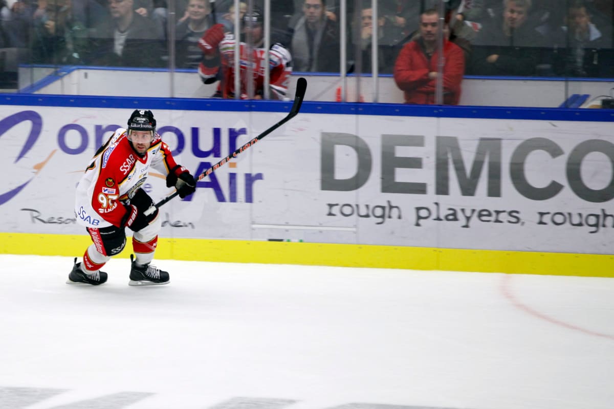 En ishockeyspelare i en röd, vit och svart matchtröja med nummer 95 och logotyper som SSAB och Läkarkjouren visas i fokus på isen. Spelaren bär full hockeyutrustning, inklusive hjälm, handskar och skridskor, håller en svart hockeyklubba och rör sig f