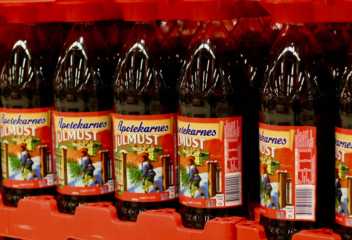Bilden visar ett antal flaskor med Apotekarnes Julmust, placerade i rött emballage eller en back. Flaskorna är mörkbruna och har röda etikettränder med texten 'Apotekarnes Julmust'. Etiketten visar även en bild av en person iklädd folkdräkt i ett snöigt vinterlandskap med granar och paketer. Det finns en streckkod synlig på en av etiketterna, samt texten 'PANT 2 KR' och '1,4 L'. Belysningen skapar reflektioner på flaskornas yta. Flaskorna är tätt packade i rad.