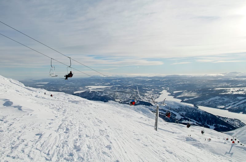 Bilden visar en vinterlandskapsvy från en skidbacke. I förgrunden syns en snötäckt bergssluttning med skidspår. Flera människor syns i skidliftar på väg uppför berget, och några figurer syns åkande skidor nerför backen. I bakgrunden sträcker sig ett