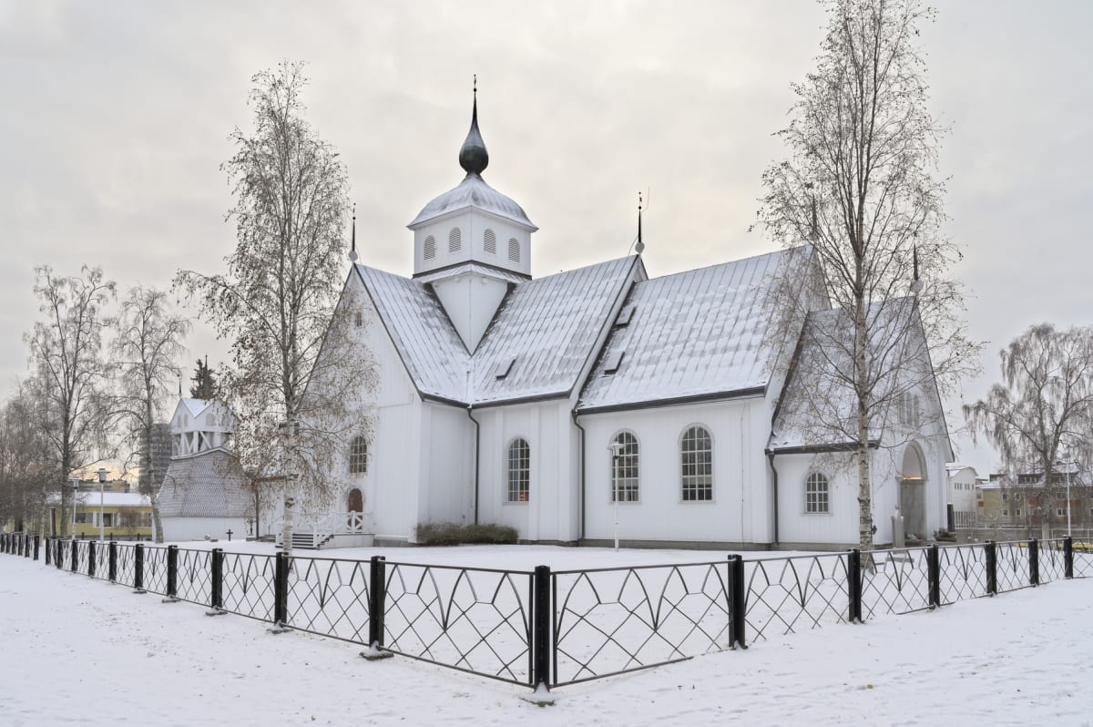 En stor vit kyrkobyggnad med snö på taket och marken. Byggnaden har en central kupolformad spira med ett ventilerat parti under. Flera sidobyggnader och takvinklar utgör kyrkans arkitektur. Runt kyrkan står kala lövträd och snötäckta buskar. Marken är täckt av ett jämnt lager snö. I förgrunden syns ett svart smidesstaket med rombformade mönster som avgränsar ett område. I bakgrunden, till vänster bortom staketet, syns en vit klockstapel och en byggnad med gula fasader. Till höger i bakgrunden syns andra byggnader belägna längre bort. Himmeln är molnig och grå.