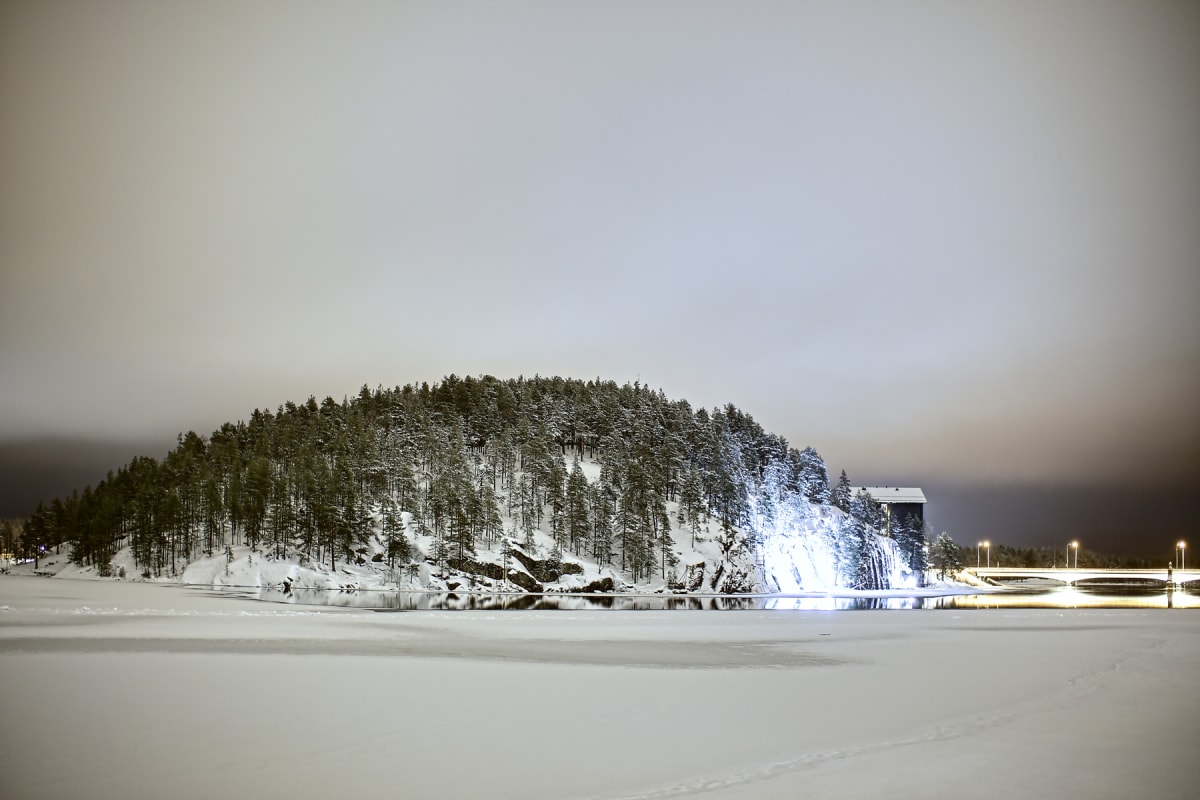 Bilden visar ett snötäckt, vitt vinterlandskap under natten eller i skymningen. I förgrunden är det ett stort, slätt område täckt av snö och is. I mitten av bilden reser sig en kulle eller bergsknall helt täckt av snötyngda granar och skog. Längs kul