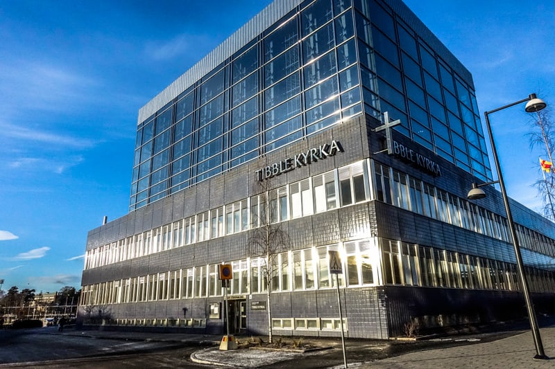 Bilden visar fasaden på en modern byggnad under dagtid. Byggnaden har flera våningar, där de övre våningarna är täckta av stora glaspartier som reflekterar himlen och solljuset. Nedre delen av byggnaden består av mörkare fasadmaterial med en mängd fö