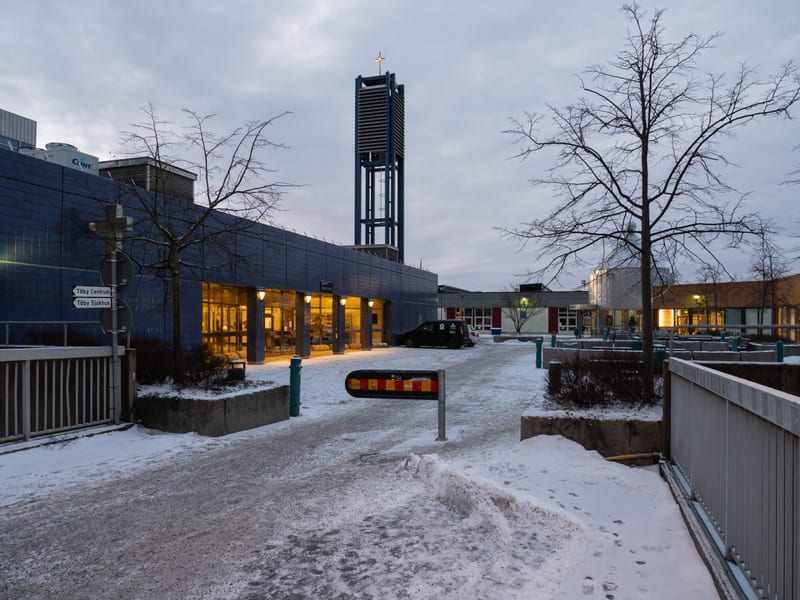 Bilden visar en vintermiljö med snö på marken och träd utan löv. I förgrunden syns en snötäckt gångväg och delar av ett vitt metallräcke till höger. Till vänster i bilden finns en vägskylt med texten "Täby Centrum" och "Täby Sjukhus" samt ett kors mo