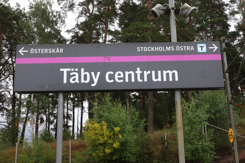 Bilden visar en stor skylt i grått med vit text och en horisontell rosa rand. På skylten står det "Täby centrum" i stor text. Ovanför "Täby centrum" finns två mindre textfält, ett till vänster med en pil som pekar åt vänster och texten "ÖSTERSKÄR", o