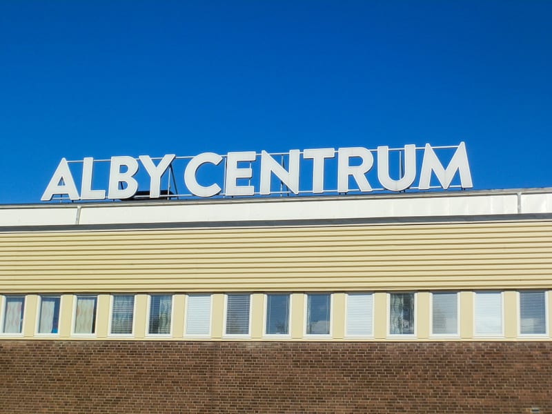 Bilden visar fasaden på en byggnad under en klarblå himmel. Överst på byggnaden står stora vita bokstäver som bildar texten "ALBY CENTRUM". Under skylten finns en ljusgul fasad med horisontella ränder, följd av en rad fönster. Fönstren är rektangulär