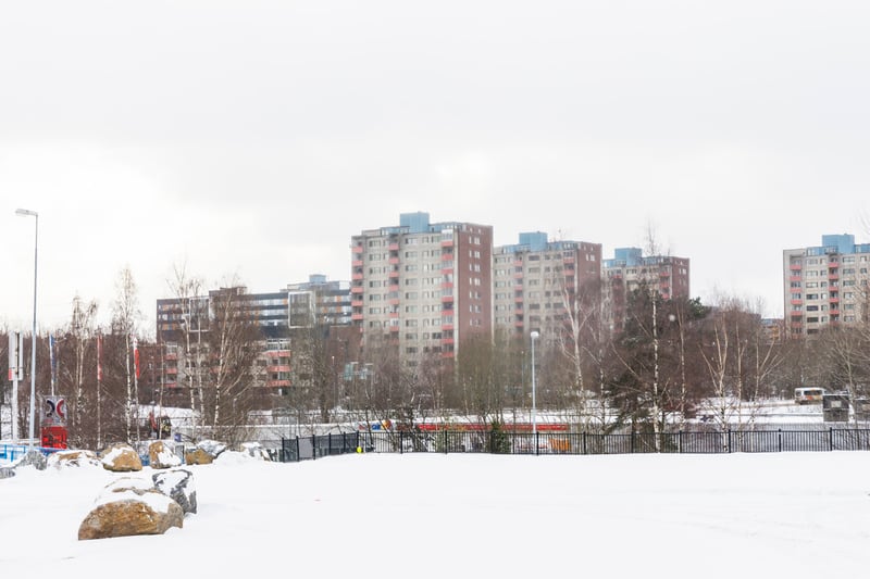 Panoramabild som visar ett snötäckt landskap med flera flerbostadshus i bakgrunden under en mulen vinterdag. I förgrunden syns snö som täcker marken, och där ligger flera stora stenar delvis övertäckta av snö. Ett svart metallstaket löper över bilden