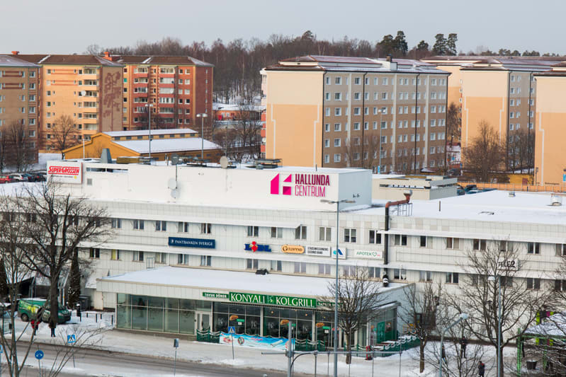 Bilden visar ett vinterlandskap med snöklädda tak och mark. I förgrunden syns Hallunda Centrum, en byggnad med flera butiksskyltar som Pantbanken Sverige, IBL, Guldfynd och Apoteket. På taket till Hallunda Centrum står en stor skylt med texten "HALLU