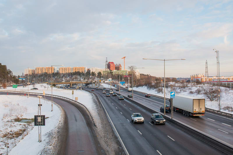 En vidvinkelbild från ovan visar en motorväg med flera filer och bilar som kör i båda riktningarna. Motorvägen har betongbarriärer och snöklädda kanter. I förgrunden syns en avfartsramp som svänger av åt vänster, också den kantad av snö. I bakgrunden