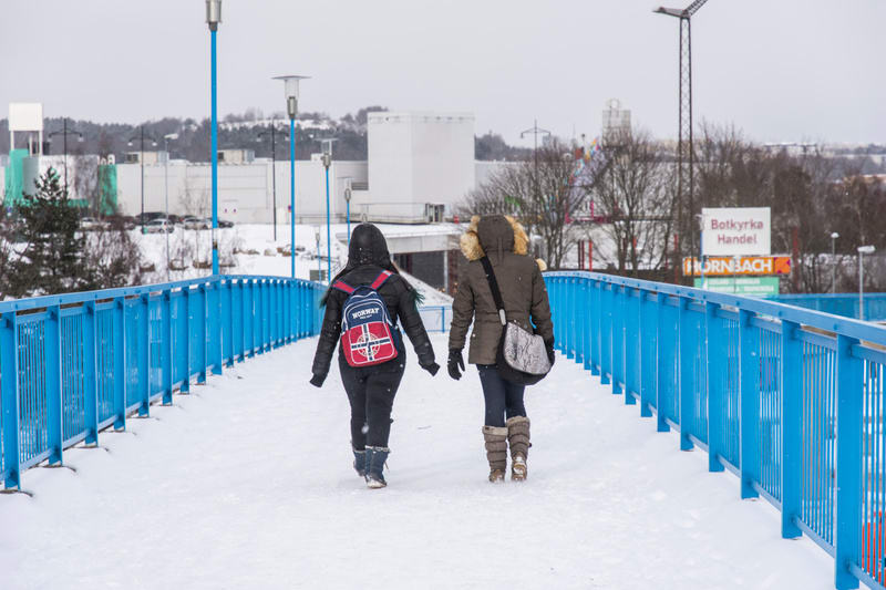 Två personer ses gå på en snötäckt gångbro med blåa räcken under snöfall. Personerna är vända bort från kameran. Den vänstra personen bär en svart jacka med luva och en ryggsäck i färgerna rött, vitt och blått med texten "NORWAY". Den högra personen