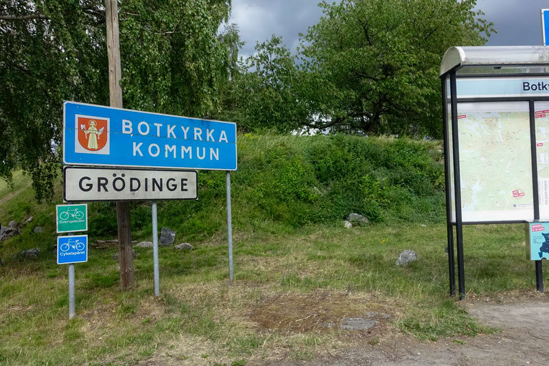 Bilden visar flera skyltar vid sidan av en väg eller stig. I centrum står en blå skylt med texten "BOTKYRKA KOMMUN" och Botkyrka kommuns vapen. Under denna skylt finns en vit skylt med texten "GRÖDINGE". Nederst till vänster syns två mindre cykelskyl