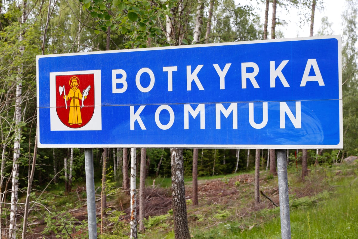 Bilden visar en blå kommunskylt med vit text som informerar om att man kommer in i Botkyrka kommun. På vänster sida av skylten finns kommunens vapen med en figur i gul klänning som håller en yxa och en fisk mot en röd bakgrund. Skylten är monterad på
