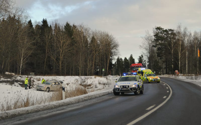 En bild tagen utomhus under dagtid som visar en olycksplats vid sidan av en asfalterad väg. I förgrunden syns en vit polisbil med blåljus aktiverade, parkerad vid vägkanten. Bakom polisbilen står en gul ambulans, även den med blåljus aktiverade. Läng