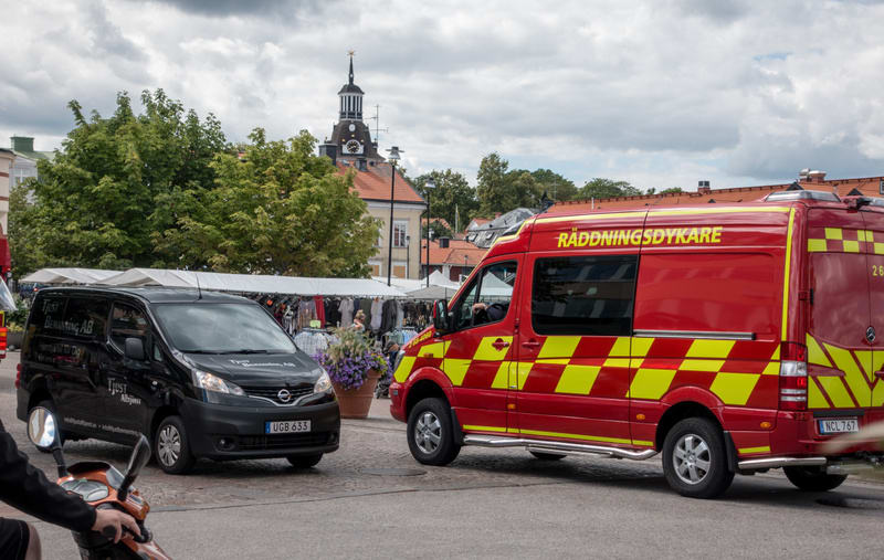 Bilden visar ett röd- och gult fordon märkt "RÄDDNINGSDYKARE" tillsammans med en svart skåpbil märkt "Tjust Bemanning AB" på en gata under en molnig himmel. I bakgrunden syns ett torg med marknadsstånd och människor. Byggnader med röda tak och grönsk