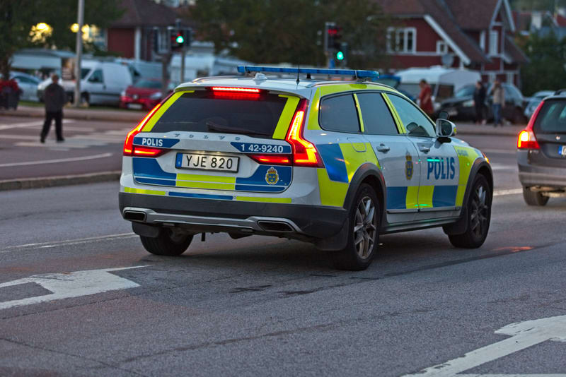 Bilden visar baksidan och vänster sida av en svensk polisbil av märket Volvo, som är ljust färgad med blå och gul ruta. Bilens baktill lyser bromsljusen. Registreringsskylten 'YJE 820' är synlig. På sidan av bilen syns texten 'POLIS' i blått. Bredvid