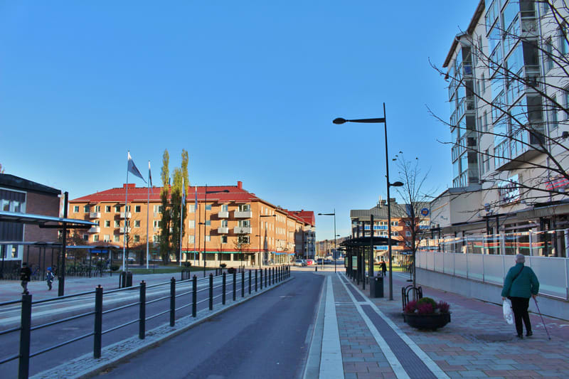 Bilden visar en gatumiljö med byggnader och öppen yta. I förgrunden till vänster finns en svart avspärrning i metall längs en asfalterad yta, som verkar vara en del av en gata eller trottoar. Till höger om avspärrningen är en markplatta med ränder so