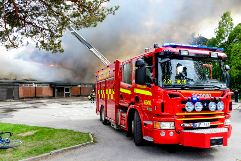 Bilden visar en röd brandbil med blåljus aktiverade, parkerad framför en byggnad med ett pågående takbrand. Byggnaden är en enplansbyggnad med mörka fasadpartier och röda tegelväggar. Tjock, mörk rök stiger upp från taket, där öppna lågor också kan s