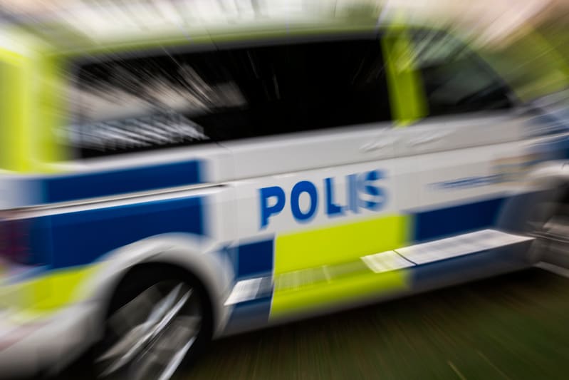 Bilden visar delen av sidan på en polisbuss med tydligt texten "POLIS" i blått. Bussen är huvudsakligen vit och har blå och limegröna (gulgröna) markeringar. Hela bilden har en effekt av rörelseoskärpa, vilket ger intryck av att fordonet rör sig snabbt. Delar av ett fordonsdäck och hjulfälgar är synliga nedtill i bilden.