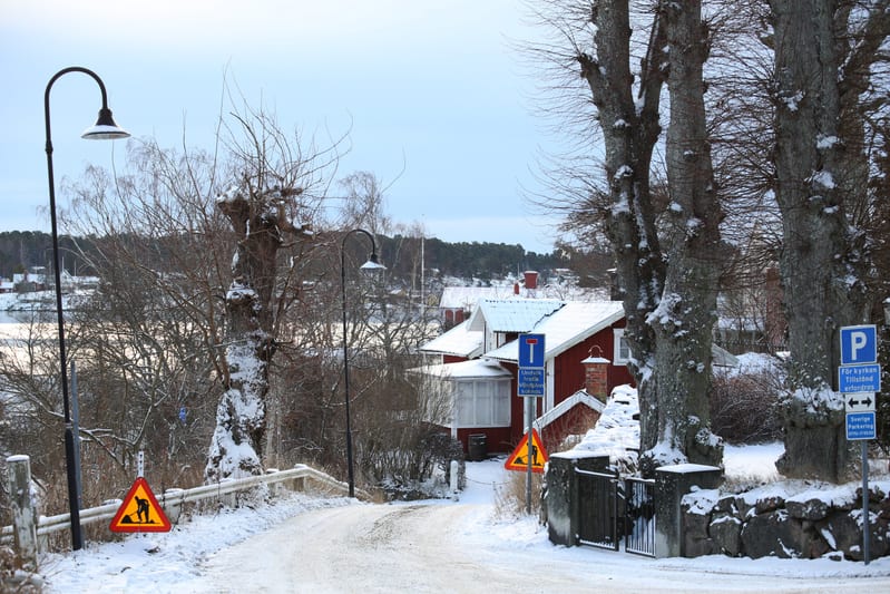 Bilden visar en snötäckt grusväg som svänger uppåt mot höger, kantad av träd och hus. På vänster sida av vägen syns en lyktstolpe och ett räcke som går längs vägen. Ett gult varningsmärke med en svart figur som arbetar syns på vänster sida av vägen. Till höger om vägen och längre fram syns flera stora trädstammar med avhuggna grenar, täckta med snö. Ett rött hus med vitt tak och vita knutar är delvis synligt mellan träden. Framför huset och till höger om vägkanten står flera skyltar; en blå ensriktad skylt med text, en blå parkeringsskylt med ytterligare text och pilar, samt ytterligare en gul varningsskylt. Marken är täckt av snö och i bakgrunden syns vatten som ser ut att vara isbelagt, omgivet av snöklädda landskap med träd och hus på avstånd. Himmelen är ljus och molnig.