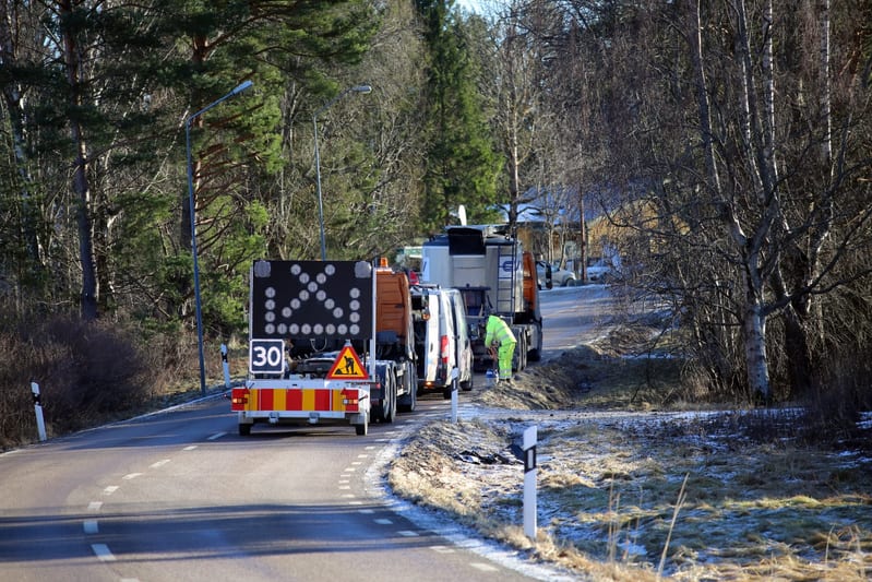 Bilden visar en väg med ett fordon utrustat med en stor varningsskylt och en hastighetsbegränsning på 30 km/h som drar en släpvagn med ett litet varningsskylt med en svart silhuett av en person som skottar jord på gul bakgrund. Vägen svänger till höger och är omgiven av träd och buskar som delvis saknar löv. Ett fordon med släpvagn är parkerat i vägkanten. En person klädd i gul arbetsjacka ses vid sidan av vägen, nära det parkerade fordonet. Markvegetationen är gräs med inslag av vissna blad och det verkar finnas spår av snö eller frost på marken. Himmelen är blå med ljusa moln, vilket indikerar dagsljus.
