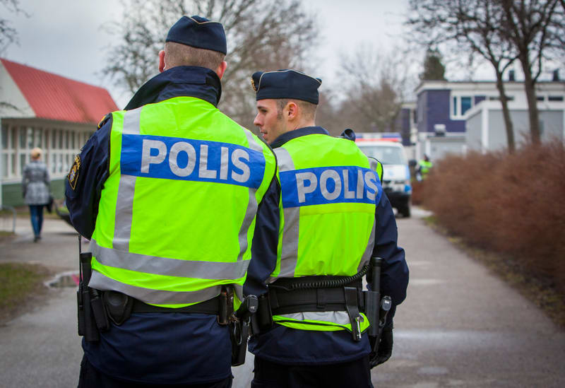 Två personer i polisuniformer, inklusive reflexvästar med texten "POLIS" på ryggen, observeras bakifrån. De bär mörkblå uniformer, mössor och har utrustningsbälten med föremål som radioapparater och batonger. Bakgrunden består av en gruskantad väg, t
