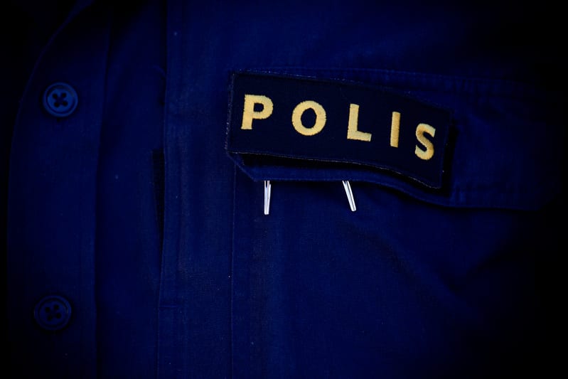 Bilden visar en närbild på en mörkblå tygjacka eller skjorta. På vänster sida av bilden syns två mörkblå knappar på en tygremsa. Jackan har en ficka på bröstet, varpå en mörkblå tygpatch med gul text som lyder "POLIS" är fäst. Under patchen, nedanför
