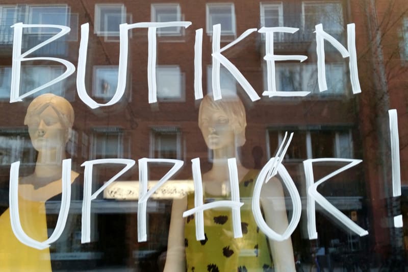 En bild tagen genom ett fönster, troligen en butiksruta, där texten "BUTIKEN UPPHÖR!" är skriven med vit färg på rutan. Bakom glaset syns två skyltdockor i förgrunden, en till vänster klädd i gult och en till höger klädd i en grönmönstrad klänning. I