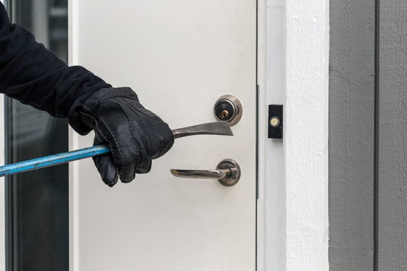 En svartklädd hand med en svart handske håller i ett blått kofotsliknande verktyg, som är placerat vid en vit dörr. Verktyget är fäst mellan dörrbladet och dörrkarmen, nära dörrhandtaget och cylindern. Bredvid dörren syns en vit dörrkarm och en mörkg