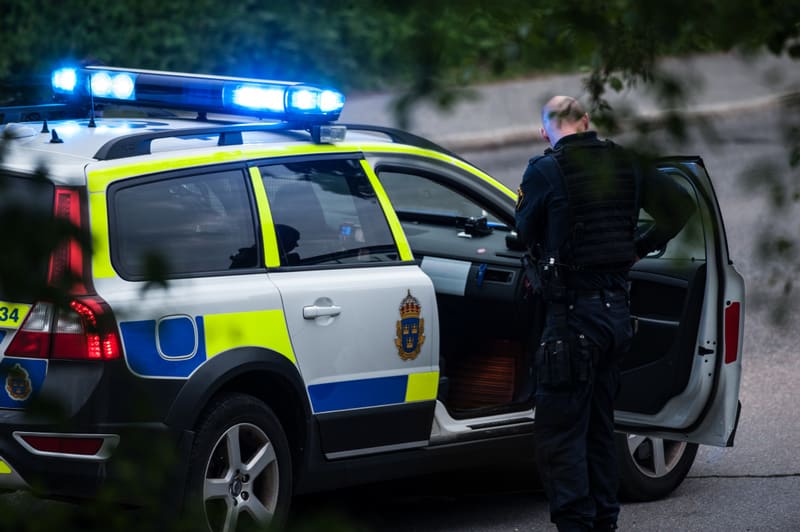 Bilden visar baksidan av en vit polisbil med blå och gul reflekterande dekor. Bilens varningsljus på taket är tända och avger ett starkt blått sken. En person, klädd i mörka kläder som ser ut att vara en uniform med utrustningsbälte och väst, står bredvid bilens förardörr vilken är öppen. Personen är vänd bort från kameran, vilket gör att ansiktet inte syns. Delar av träd eller buskar, med grönt lövverk, skymtar i förgrunden och bakgrunden, vilket indikerar att bilden är tagen utomhus.