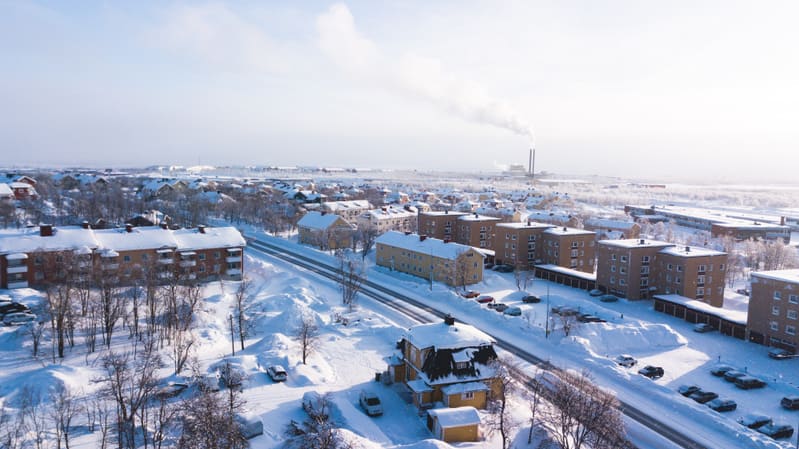 En vidvinkelbild tagen från ovan, som visar ett vinterlandskap täckt av snö. I förgrunden och mitten syns flera bostadshus, både flerfamiljshus och fristående villor, med snötäckta tak. Mellan byggnaderna löper vägar som också är snötäckta, men med s