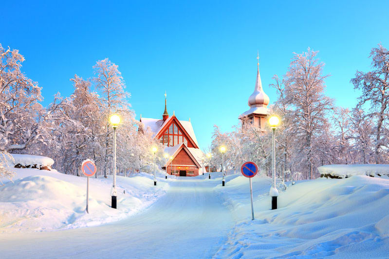 Bilden visar ett snötäckt vinterlandskap med en byggnad som liknar en kyrka i förgrunden. Byggnaden har rött tak och träfasad med en spetsig tornspira i mitten och en kupolformad sidospira till höger, båda täckta med snö. En snötäckt väg leder upp mo