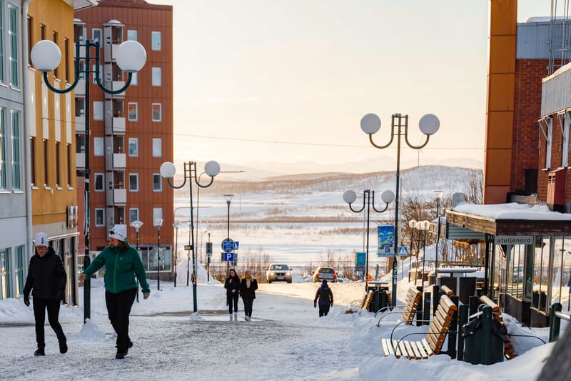 Bilden visar en snötäckt gata i en stadsmiljö under vad som verkar vara sen eftermiddag eller tidig morgon med ett ljust, gulaktigt sken på himlen. Flera personer, klädda i vinterkläder, promenerar på gatan som delvis är plogad men fortfarande har sn