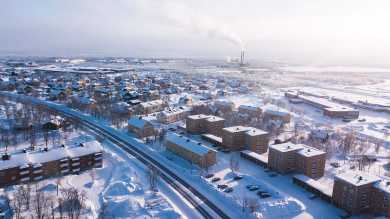 En vidvinkelbild tagen uppifrån över ett snötäckt bostadsområde under vintertid. Bilden visar flera flerbostadshus med gul-bruna fasader och rödbruna tegeltak, täckta med ett tjockt lager snö. Mindre villor av varierande färg, även de snötäckta, syns