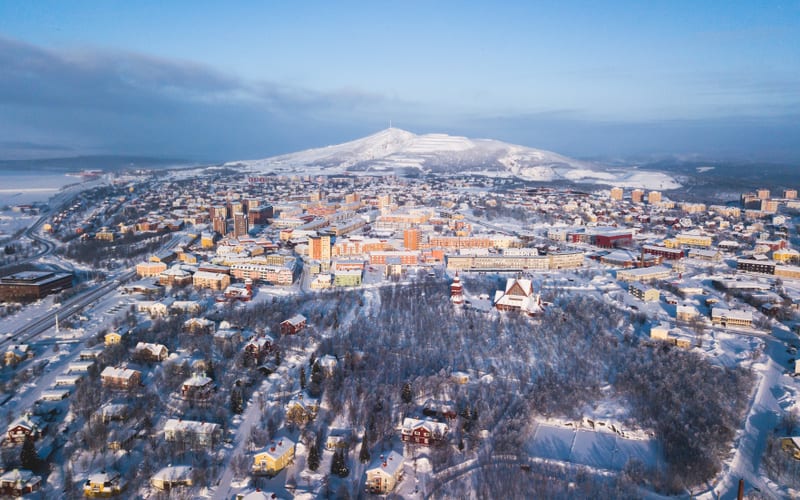Denna flygbild visar en stad och dess omgivningar täckta av snö under dagtid. I förgrunden syns ett skogsområde med snötäckta träd och flera bostadshus med snö på taken. Upp till höger i bilden avtecknar sig en kyrkobyggnad med ett rött tak och en sp