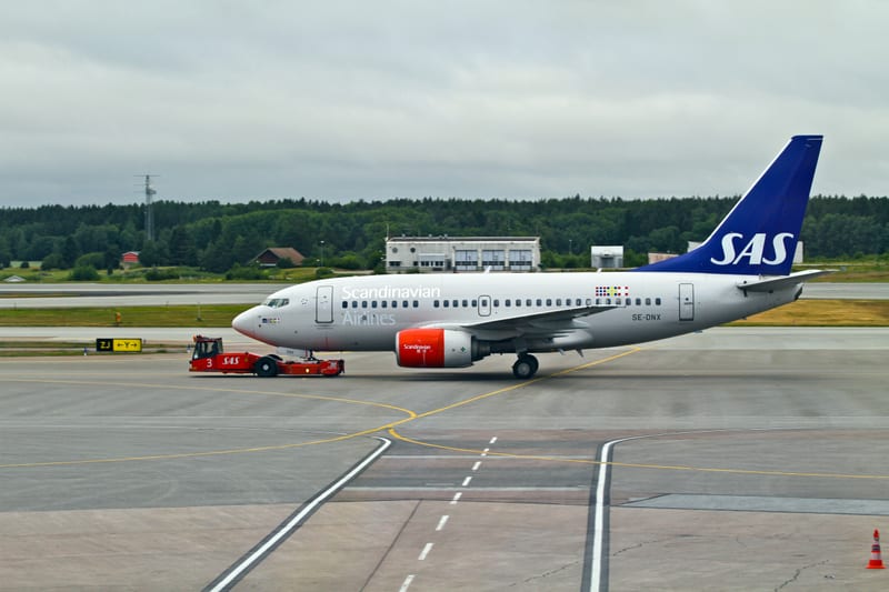 En bild visar ett flygplan från Scandinavian Airlines (SAS) med registreringsnummer SE-DNX på en landningsbana eller taxibana. Flygplanet är vitt med blå detaljer på stjärtfenan och texten "SAS" i vitt. Motorn på ena sidan är delvis synlig och har en orange färgton. En röd flygplansdragare, med texten "SAS" på sidan, är fäst vid flygplanets noshjul. I bakgrunden syns skog, några byggnader och en mast under en gråmulen himmel. Markeringar på asfaltsytan är också synliga.