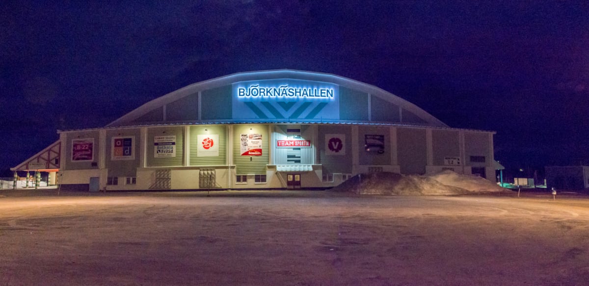 En fasadbild på en stor byggnad med texten "BJÖRKNÄSHALLEN" upplyst på taket. Byggnaden är belyst med skyltbelysning längs fasaden som framhäver flera rektangulära annonstavlor och logotyper. Till vänster syns delar av en sidobyggnad med ett takstati