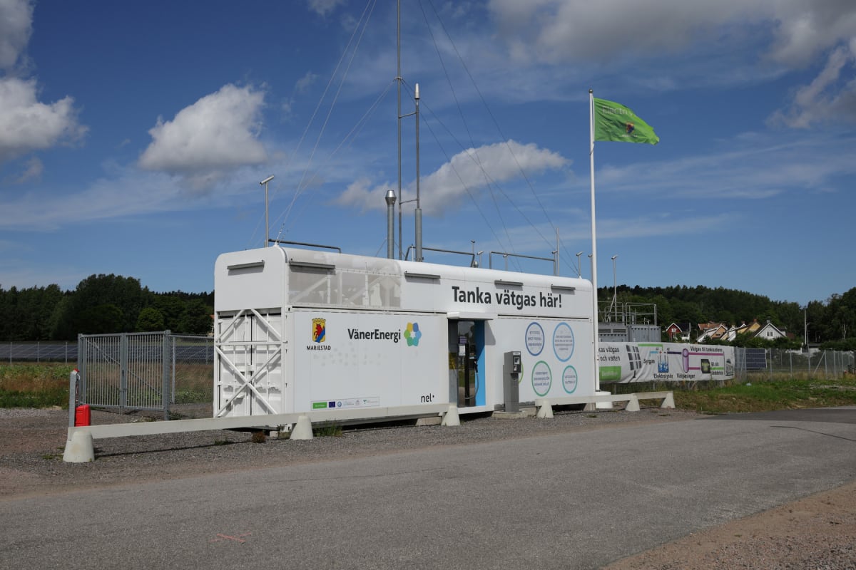 En modern vätgasstation står placerad utomhus under en klarblå himmel med spridda moln. Stationen består av en stor vit containerliknande byggnad med texten "Tanka vätgas här!" på sidan. Flera skyltar och logotyper såsom "VänerEnergi" och "Mariestad"