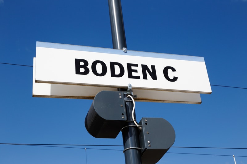Bilden visar en vit skylt med texten "BODEN C" i svarta versaler, monterad horisontellt på en mörk stolpe. Under skylten, på samma stolpe, sitter två mörka, rektangulära högtalarliknande anordningar, fästade med metallbeslag. En vit kabel är lindad r