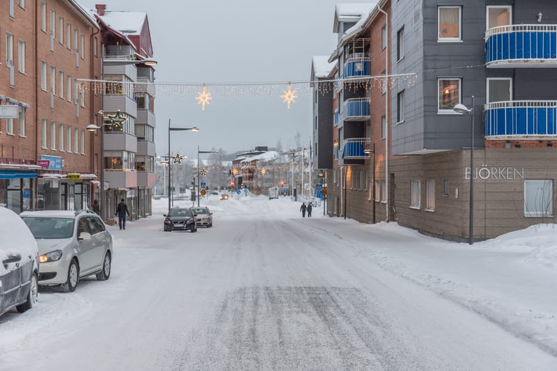 En ganska bred stadsgata är täckt av snö, med snövallar längs sidorna vid trottoarkanterna. På båda sidor av gatan finns flervåningsbyggnader med lägenheter och butiker på markplan. Vissa butiksfasader har skyltfönster med skyltning. Bilar står parke