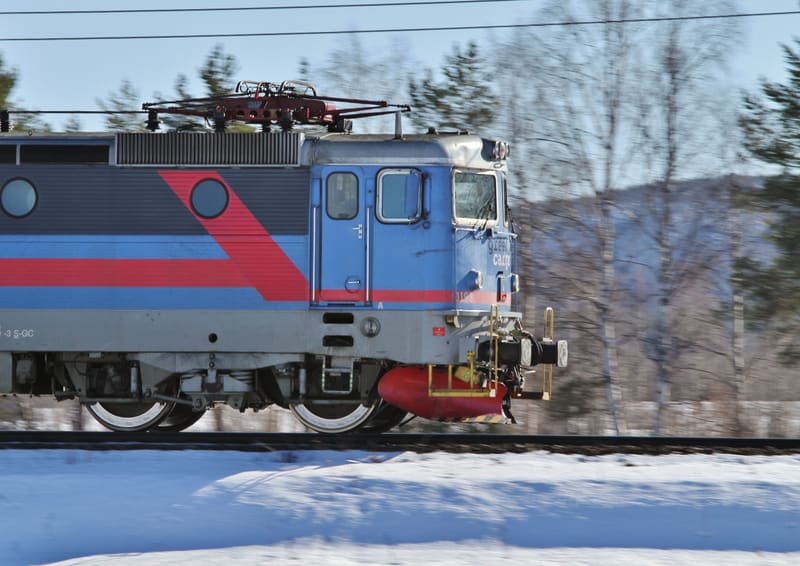 Bilden visar en blå, grå och röd lokomotiv för gods under rörelse i ett snötäckt landskap med träd i bakgrunden. En mindre del av lokomotivets främre del är synlig och har texten "Green Cargo" på sidan. Flera fönster kan ses på lokomotivet, samt deta