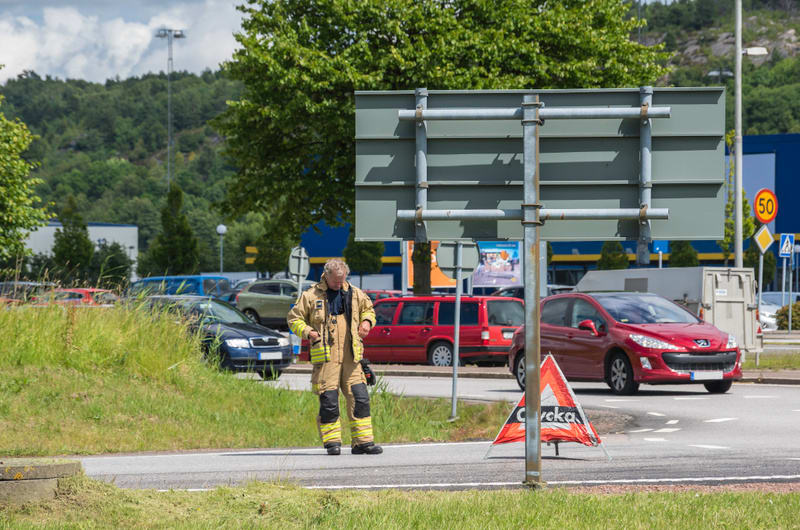 En man klädd i brandmansutrustning med gulaktiga byxor och jacka står på en väg eller parkering. I bakgrunden syns flera parkerade bilar i olika färger, bland annat en röd kombi, en mörkblå sedan och en röd hatchback. En grön grässlätt med högt gräs