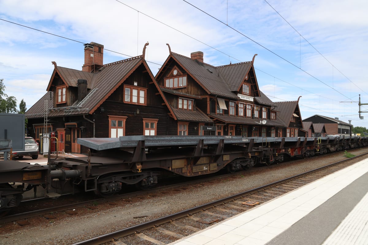 En bild som visar en järnvägsstation med ett mörkt träfärgat stationshus med bruna tak och många fönster. Stationshuset pryds av gaveldekorationer och har en skylt med texten "BODEN CENTRAL". Framför stationshuset står flera godsvagnar på järnvägsspå