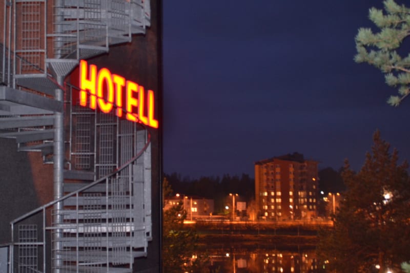 Bilden visar fasaden på en byggnad med ett spiraltrapphus och en röd neonljusskylt där det står ”HOTELL” i versaler. Skylten är belyst och trapphuset är tillverkat av vad som verkar vara metall. I bakgrunden syns en mörkblå natthimmel och bebyggelse