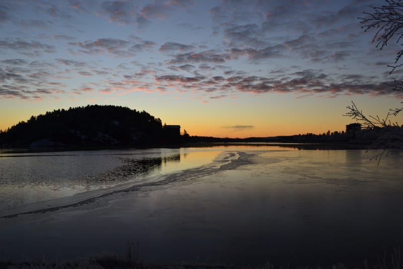 Bilden visar ett landskap med en klar himmel som skiftar från orange vid horisonten till blått högre upp, med strimmor av vita moln som reflekterar rosa nyanser. I mitten av bilden ligger en sjö eller större vattenyta som delvis är täckt av is, med e